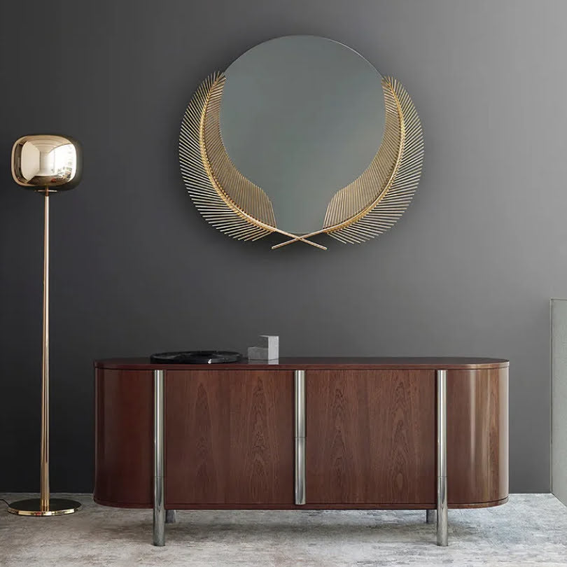 A luxurious round mirror with gold palm-leaf accents, styled in an elegant interior with a dark wall, wooden sideboard, and floor lamp.