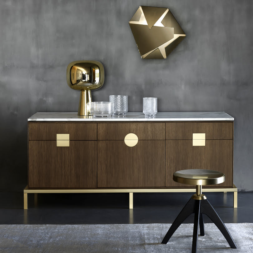 A luxurious wooden sideboard with brass inlays, topped with decorative objects, against a moody grey wall with geometric gold wall art.