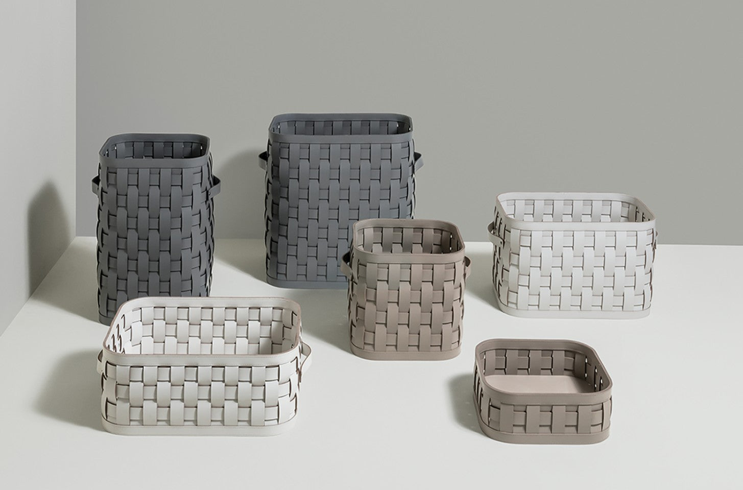 A collection of woven storage baskets in neutral grey, taupe, and white tones, arranged in two rows against a minimalist background.