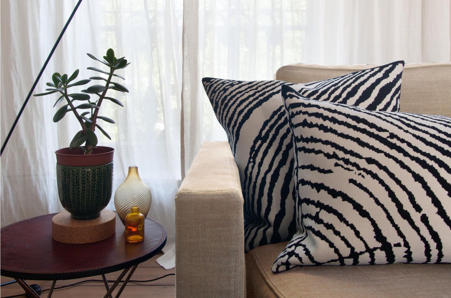 A cozy living room corner with a beige sofa, black-and-white patterned throw pillows, a round side table holding a potted plant and decorative glass vases.