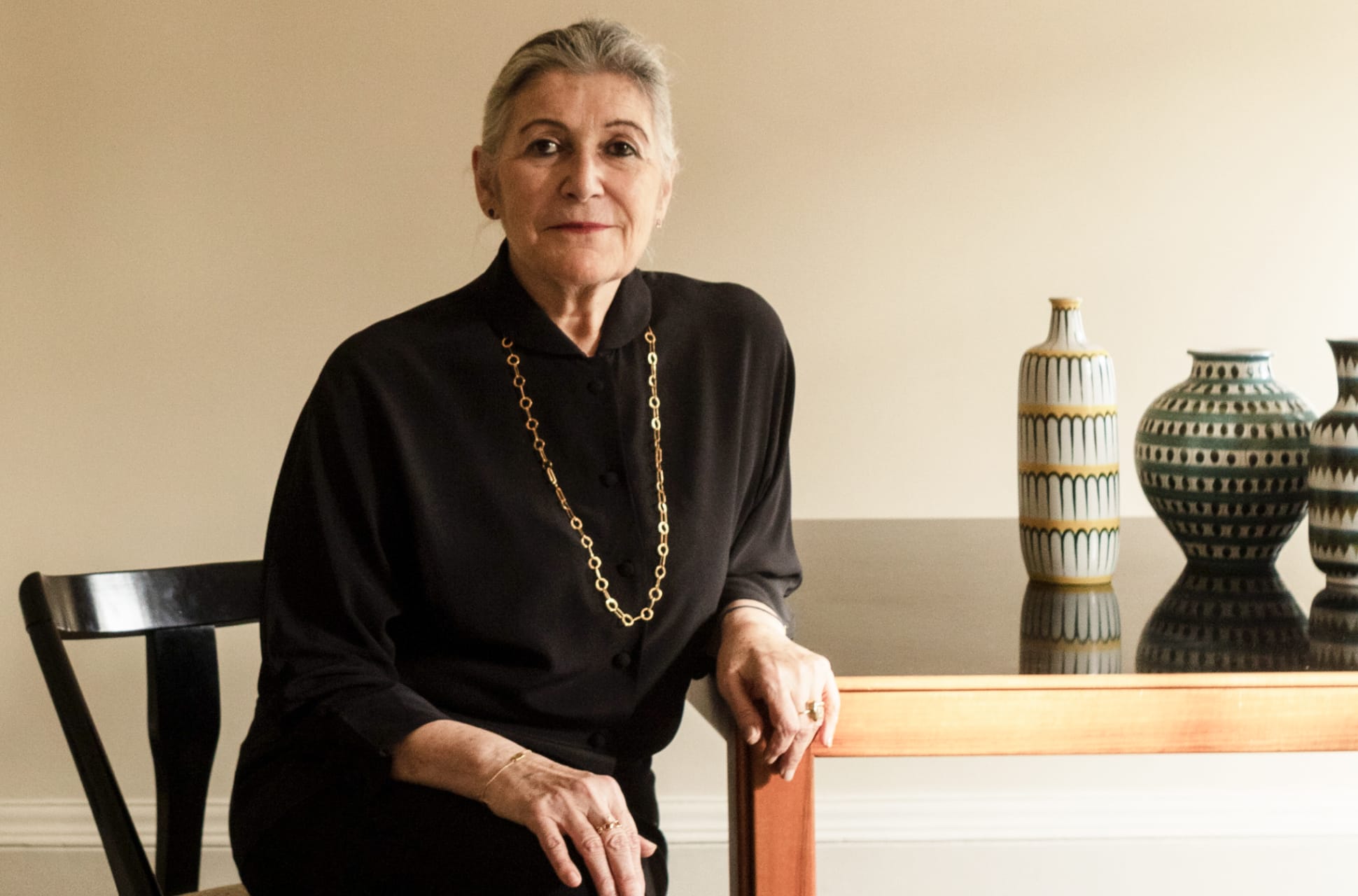 Portrait of textile designer Christine Van Der Hurd seated beside a table, wearing a black outfit and gold chain necklace.