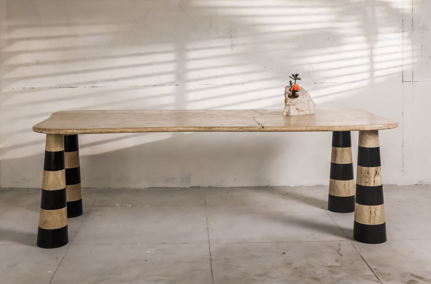 A sculptural travertine table with bold striped conical legs, set against a textured wall with soft shadow patterns, blending artistry with functional design.