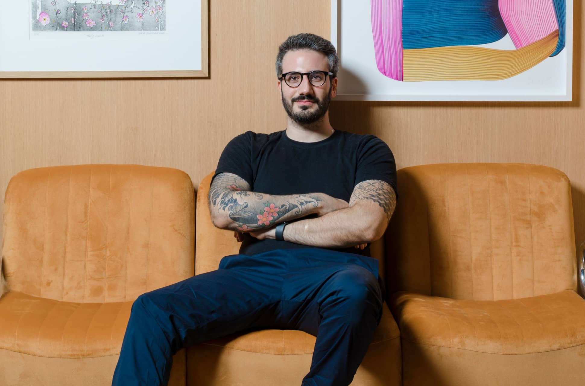 Designer Thomas Trad seated on a sculptural mustard velvet sofa with scalloped edges, wearing a black t-shirt, navy trousers, and white sneakers, with arms crossed and a confident, relaxed demeanor, set against a backdrop of contemporary framed artworks.