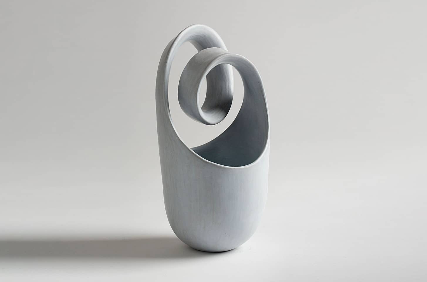 A sculptural ceramic vessel by Tina Vlassopulos in a soft grey tone, featuring an elongated, elliptical body with a continuous looping form rising from its rim, exemplifying fluidity and balance.