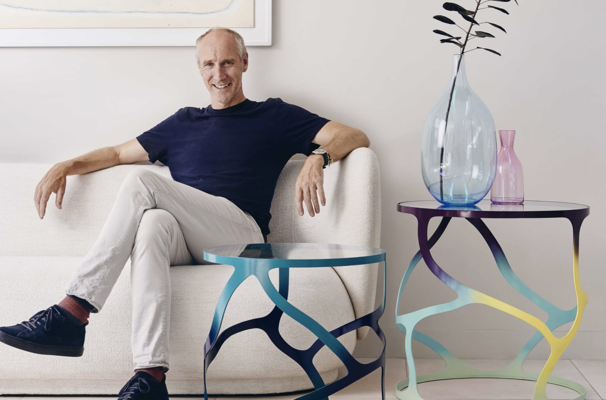 Designer Tom Faulkner seated on a modern cream-colored sofa, wearing a navy t-shirt and light trousers, with a contemporary sculptural side table in front of him featuring a blue, organic cut-out metal base and a glass top.