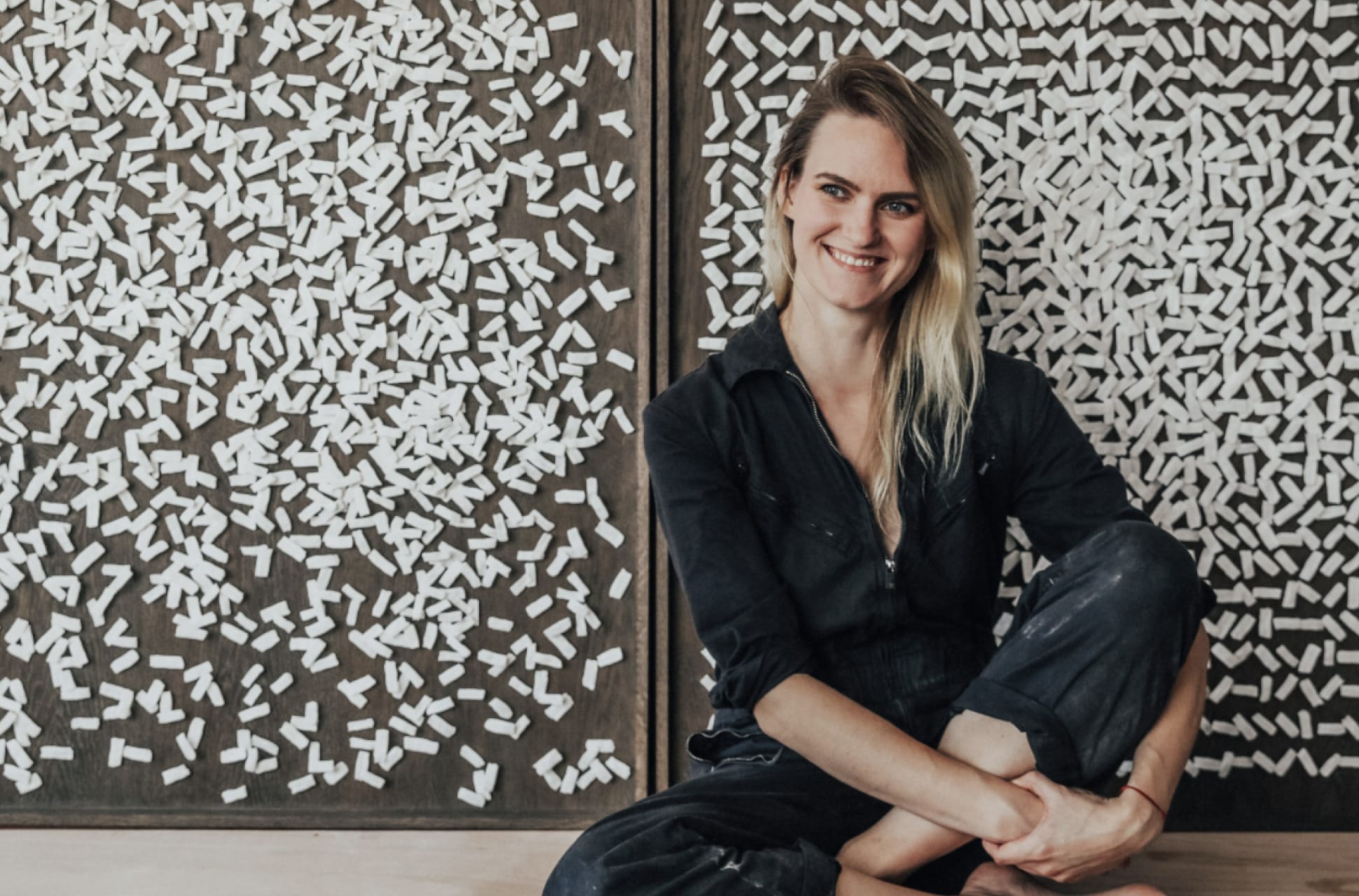 Ula Saniawa sitting on the floor in front of a textured wall with scattered white shapes, wearing a dark jumpsuit and smiling.