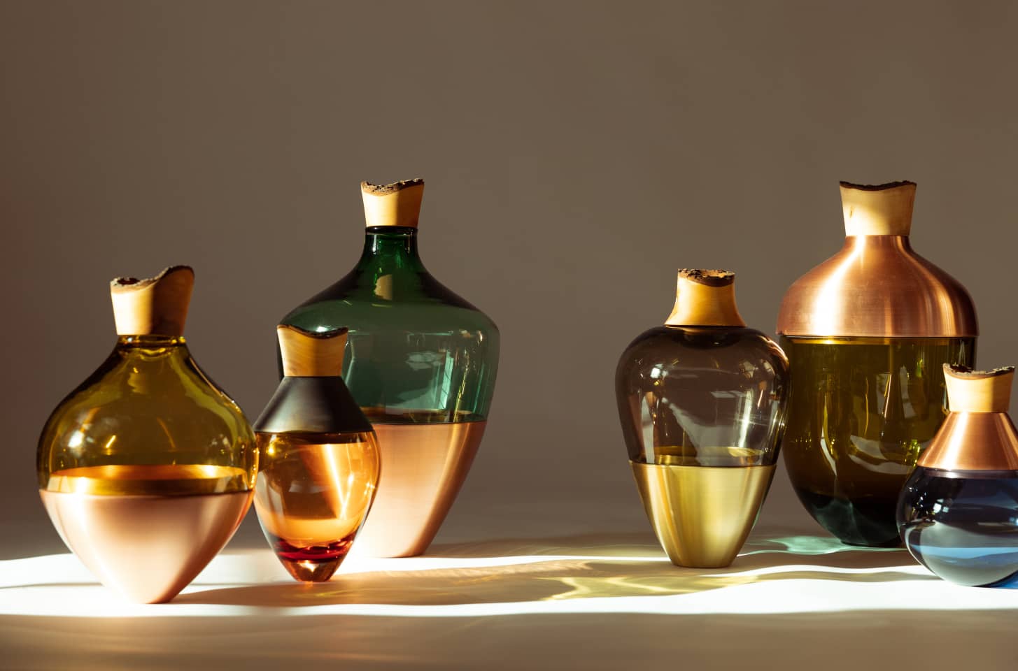 Glass vessels in various colors with copper and wood tops, arranged in warm light.