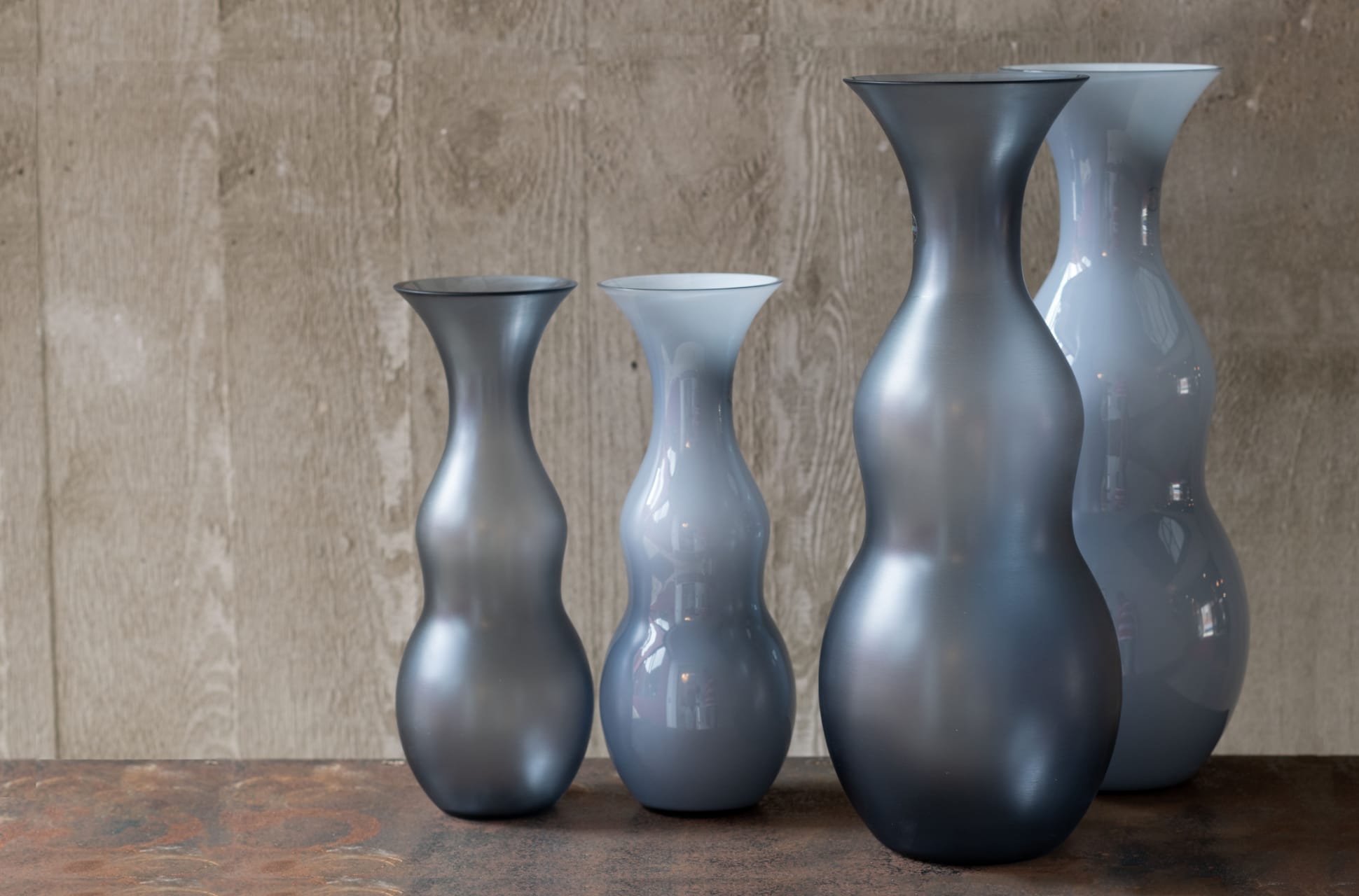 Four curvaceous glass vases in varying heights and shades of smoky blue and grey, displayed on a textured surface against a wood-panel background.