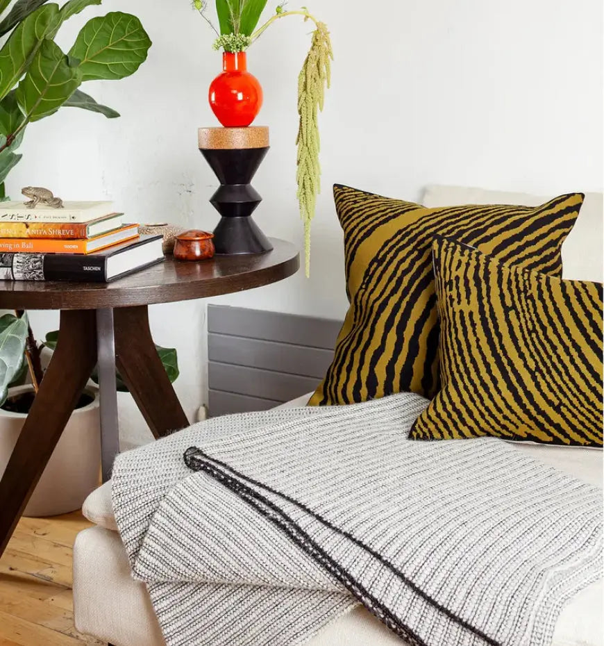 Corner of a sofa with striped pillows, a textured blanket, and a wooden table holding books and a vibrant red vase.